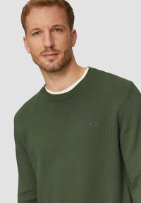 Olivengrüner gestrickter Pullover mit Rundhalsausschnitt und geripptem Material. Mit kleinem Markenlogo auf der Brust und weißem Detail am inneren Kragen.
