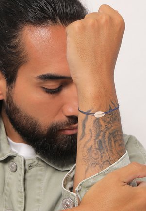 Blå snorarmbånd med et lille, hvidt muslingebeslag, båret på en underarm med indviklede tatoveringsmønstre, mod en neutral baggrund.