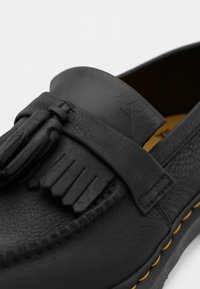 Detailný záber na čierne kožené loafery s strapcami a žltým prešívaním pozdĺž podrážky.