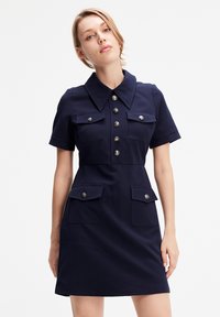 Robe courte noire à manches courtes avec un col, un devant boutonné, deux poches poitrine et deux poches à rabat inférieures, fabriquée dans un tissu lisse.