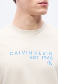 Κοντινό πλάνο ενός ατόμου που φοράει ανοιχτό μπεζ μπλουζάκι με μπλε κείμενο «Calvin Klein EST. 1968 cK» στο στήθος.