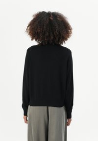 Pull noir en tricot avec un col rond et des manches longues, doté d'un ourlet côtelé. Le modèle porte un pantalon large gris clair.