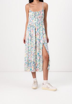 Femme portant une robe midi fleurie pastel avec fines bretelles et fente latérale, assortie à des chaussettes blanches et des baskets, debout devant un fond blanc.