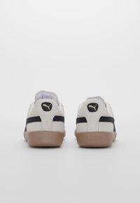 Sneakers bianchi in suede con una striscia nera e suola in gomma beige. Sul retro è visibile un piccolo logo dorato e un collarino imbottito.