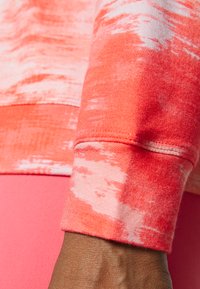 Tecido de sweatshirt cor-de-rosa com um padrão de tie-dye, apresentando um punho ajustado com detalhes em costura e uma textura suave. Parte de uma peça de vestuário rosa visível.