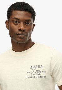 Camiseta de manga corta color crema con cuello redondo. Presenta un logo impreso en gris en el pecho, con una textura suave.