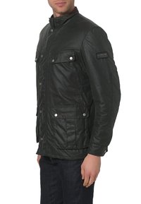 Donkergroene gewaxte jas met een hoge kraag, drukknoopsluitingen, twee voorzakken en een zijlogo-patch, met een gestructureerd oppervlak.