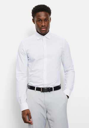 Man wearing a fitted white button-up shirt tucked into light gray pants with a black belt, standing with one hand in pocket.