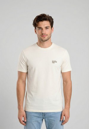 Giovane in piedi, indossa una T-shirt a maniche corte color crema chiaro con la scritta piccola "Armani Exchange Club" sul petto sinistro e jeans azzurri.