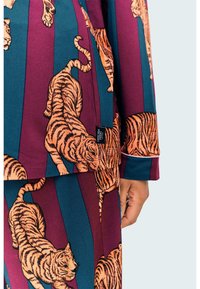 Conjunto de pijama con un patrón multicolor de tigres naranjas sobre rayas burdeos y verde azulado, con una etiqueta de tela visible en el dobladillo.
