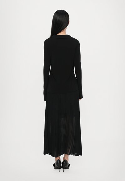 Black pleated dress featuring long sleeves and a collar, with a flowy skirt and split detail at the back, paired with black high-heeled shoes.