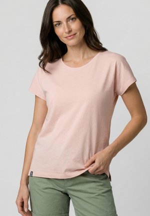 Frau mit braunen Haaren, die ein hellrosa kurzärmeliges Shirt und grüne Shorts trägt, hält den Saum des Shirts mit einer Hand fest.