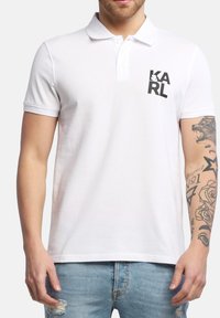 Polo bianco in cotone, con colletto classico, maniche corte e un logo stampato nero sul lato sinistro del petto. Visibili jeans in denim.
