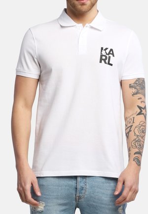 Hvid polo-shirt lavet af bomuld, med en klassisk krave, korte ærmer og et sort trykt logo på venstre bryst. Denim jeans synlige.