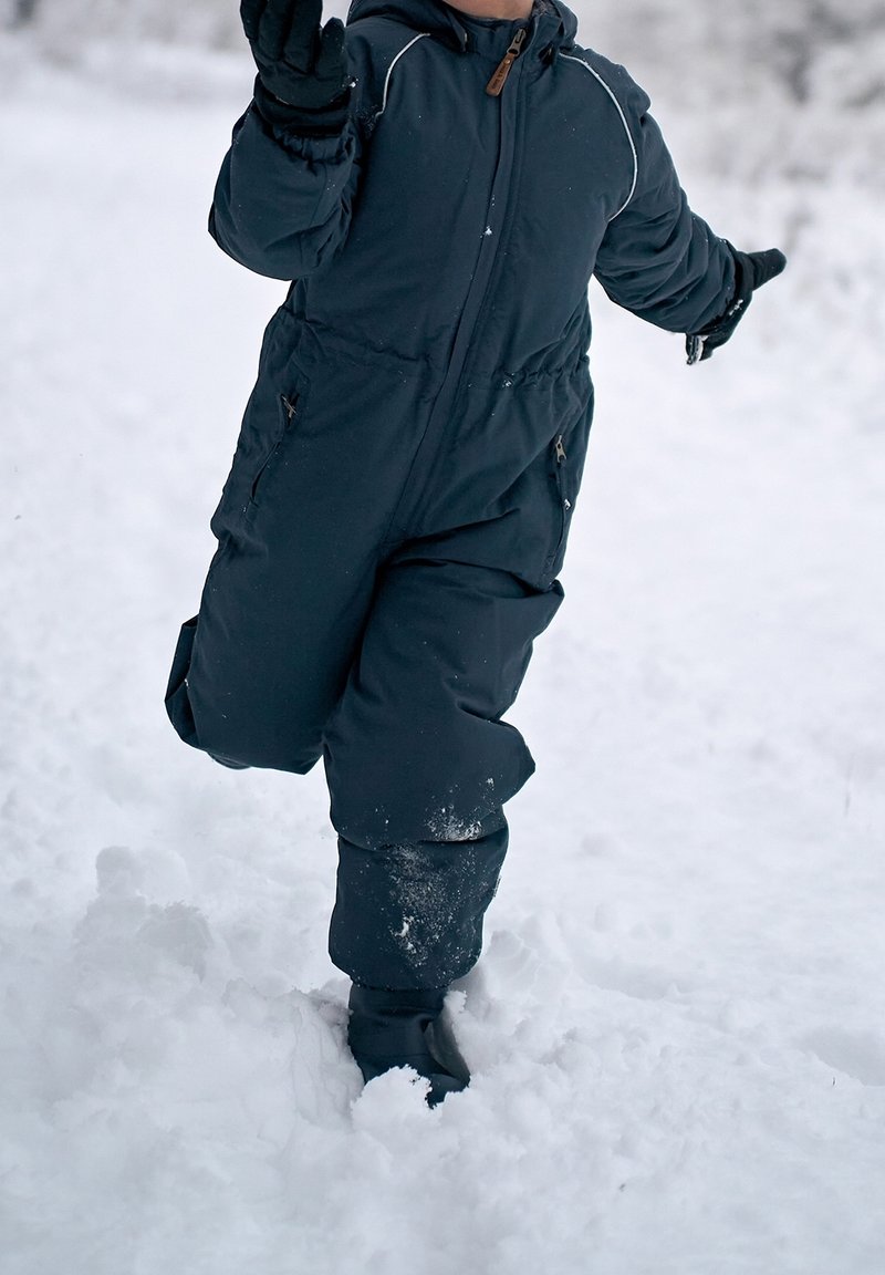 Enfant en combinaison de neige sombre et gants marchant dans une neige profonde à l'extérieur, se tenant en équilibre les bras tendus.