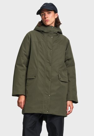 Personne portant un long manteau à capuche vert olive avec des boutons-pression et des poches à rabat, associé à une casquette noire et un pantalon noir.