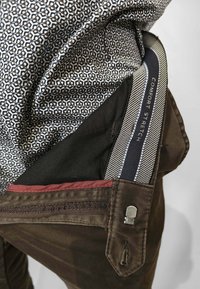 Pantaloni in cotone marrone con chiusura a zip e dettagli. Presentano una cintura elasticizzata a righe con l'etichetta "Comfort Stretch" e un tessuto con motivo.