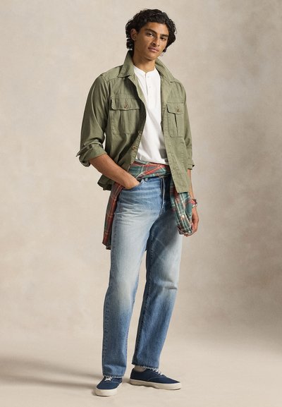 Χακί μιλιτέρ μπουφάν πάνω από λευκό πουκάμισο henley, ανοιχτό μπλε παντελόνι ίσιας γραμμής, και μπλε αθλητικά παπούτσια, με ένα καρό πουκάμισο δεμένο στη μέση.