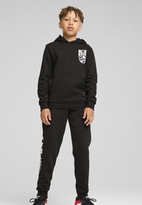 Juodas treningas su priekiu kišene, logotipu ant krūtinės ir derančiais juodais kelniais. Medžiaga turi lygų paviršių, su elastingais rankovėmis ir liemeniu.