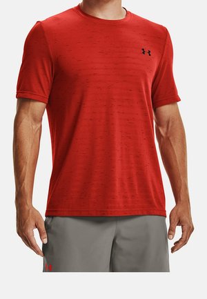 Mężczyzna w czerwonej sportowej koszulce z krótkim rękawem z małym czarnym logo Under Armour oraz szarych szortach z czerwonym logo.
