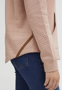 Sweatshirt rose clair avec petits pois bordeaux, doté d'une poche et d'un accent marron à l'ourlet. Porté avec un jean bleu foncé.