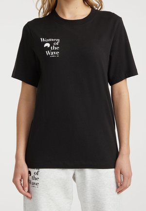 Person iført en sort T-shirt med teksten "Women of the Wave" og hvide joggingbukser med matchende tekst på venstre lår.