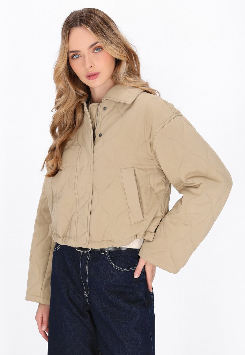 Afskåret quiltet jakke i beige stof med krave, trykknapper og sidelommer. Sammen med mørke denimbukser.