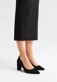 Czarne zamszowe buty na wysokim obcasie z wydłużonym noskiem, masywnym obcasem i wycięciami po bokach. Gładka faktura i elegancki design.