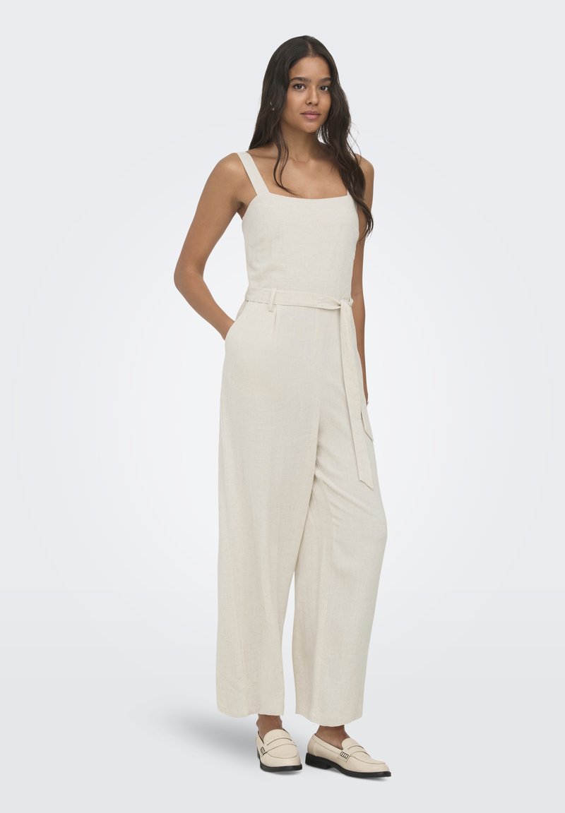 Femme aux longs cheveux foncés portant une combinaison beige sans manches avec ceinture et des mocassins beiges, debout avec une main dans la poche.