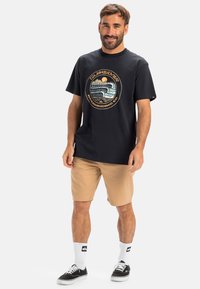 Black cotton t-shirt featuring a circular logo in earthy tones. Paired with beige shorts and black sneakers, with white crew socks.