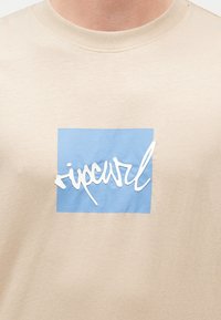 Μπεζ μπλούζα με μπλε τετράγωνο και λευκή καλλιγραφική επιγραφή "ripcurl" στο κέντρο του στήθους.