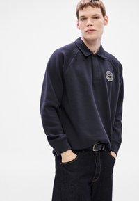 Μπλε ναυτικό πουλόβερ με γιακά, διακοσμημένο με στρογγυλή λογότυπη τσέπη, μακριά μανίκια και ριμπ μανσέτες. Φορεμένο πάνω από σκούρο τζιν.