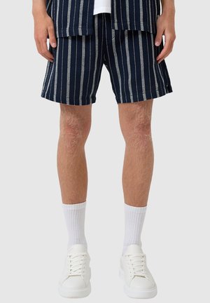 Beine mit marineblauen Shorts mit weißen vertikalen Streifen, weißen Socken bis zur Mitte der Wade und weißen Turnschuhen vor einem einfarbigen Hintergrund.