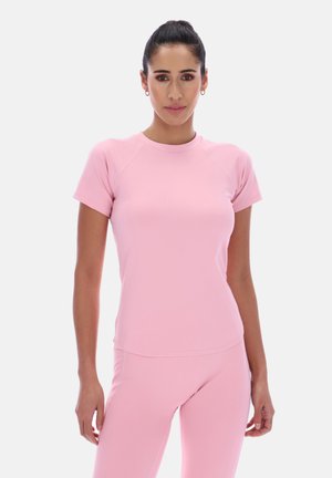 Femeie în picioare, purtând un top roz deschis cu mâneci scurte, mulat, și pantaloni roz asortați, pe un fundal simplu, deschis.