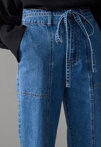 Blå denim høy midje bukser med knyttet midje, med sidelommer og en jevn tekstur. Søm detaljer langs sømmene.