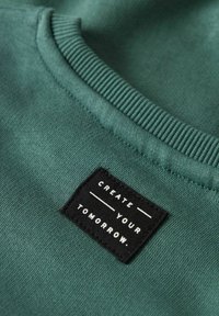 Zielony materiał z prążkowanym kołnierzykiem oraz czarną prostokątną naszywką z białym napisem "STWÓRZ SWOJĄ PRZYSZŁOŚĆ.".