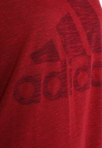 Punainen puuvillapaita, jossa on teksturoitu, haalistunut printti Adidas-logosta tummemmissa punaisissa sävyissä. Lyhyet hihat, pyöreä kaula-aukko.