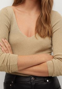 Haut à manches longues en maille dorée côtelée, col rond, tissu texturé, coupe ajustée et manches retroussées, associé à un pantalon en cuir noir.