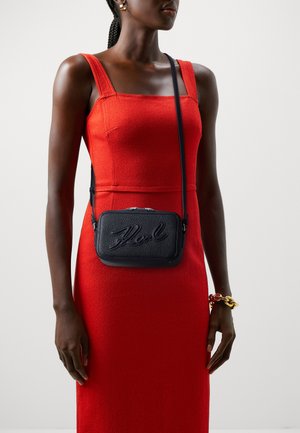 Borsa a tracolla nera con texture, logo impresso, dotata di chiusura con zip e tracolla regolabile, indossata sopra un vestito rosso aderente.