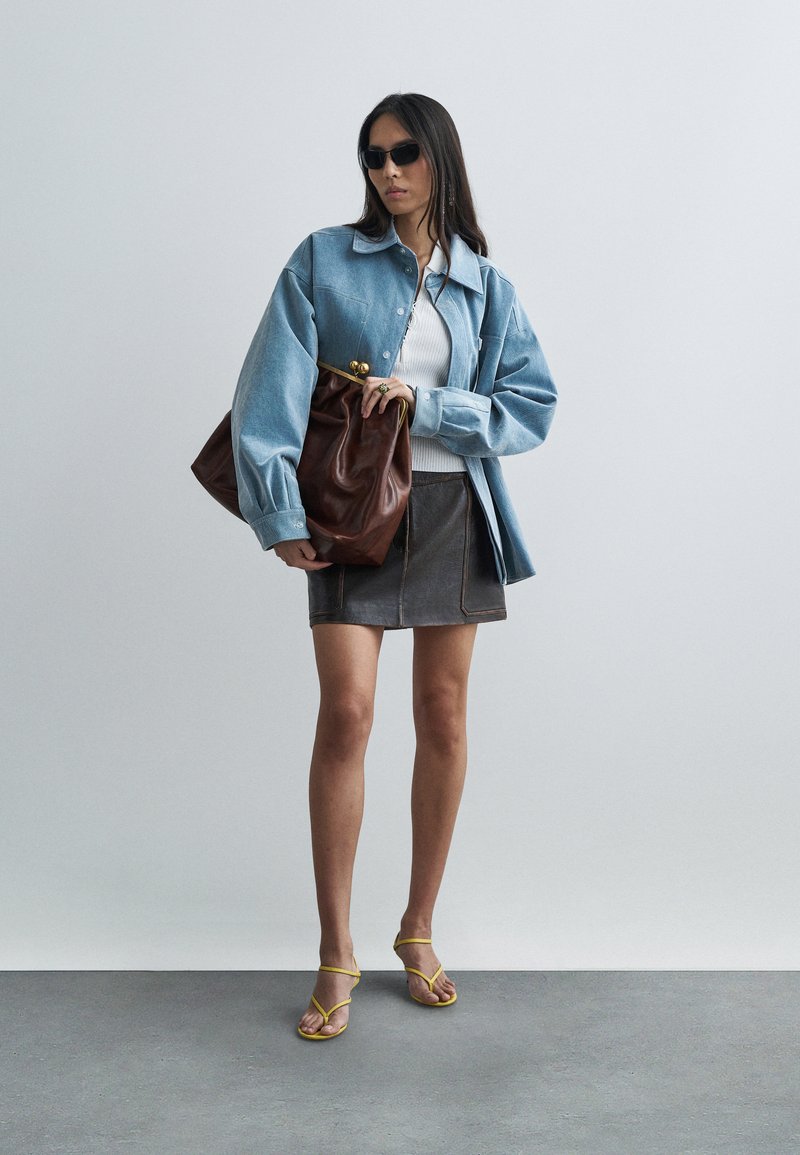 Woman wearing oversized blue denim jacket, white top, dark leather mini skirt, yellow sandals, sunglasses, holding large brown leather clutch bag.