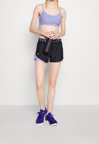 Lila sportbh med strukturerade ränder, svarta träningsshorts med ljuslila detaljer och mörklila sneakers. Håller i en svart vattenflaska.