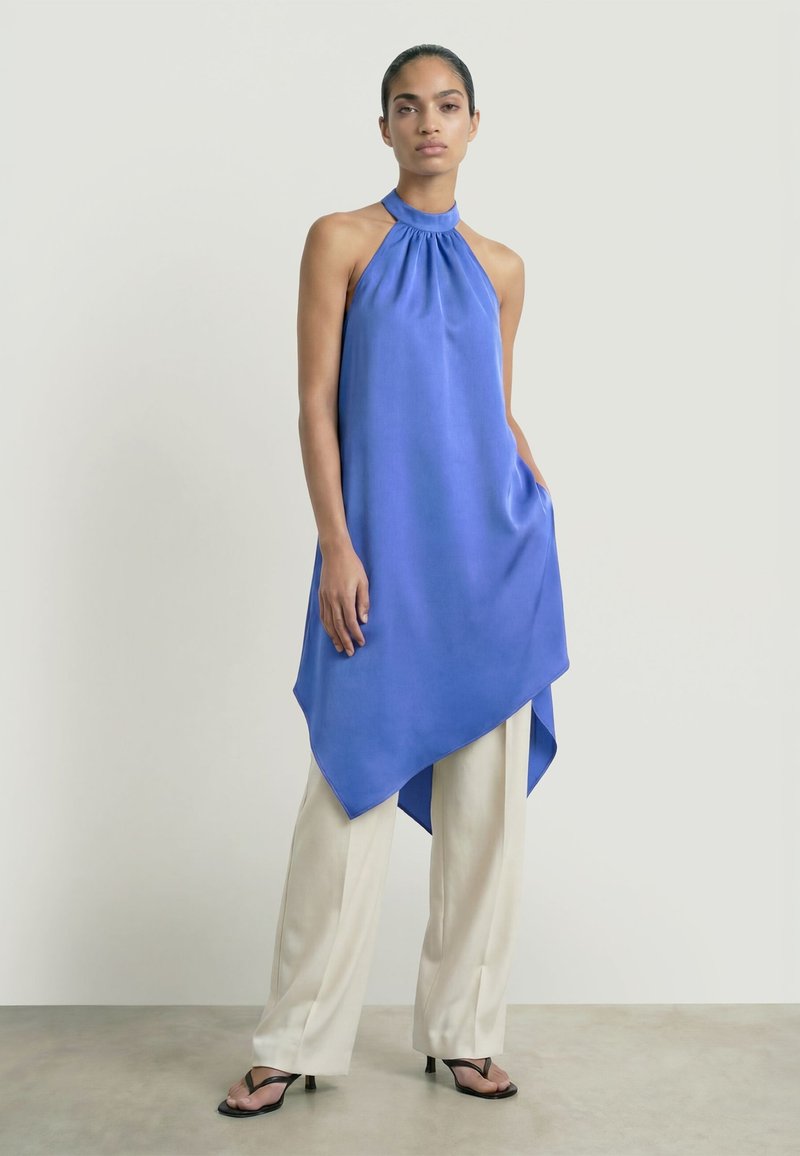 Femme portant un haut sans manches bleu à col licou avec un ourlet asymétrique sur un pantalon blanc à jambes larges et des talons ouverts noirs, se tenant devant un fond uni.