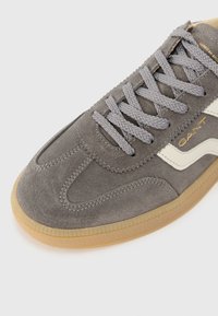 Pilkos suede sportbačiai su gumuotu rubberiniu padu, pilkomis raištelėmis ir balta juostele. Yra siuvimo detalės ir auksinė logo šone.