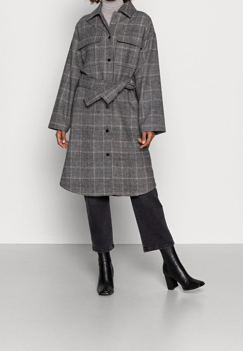 Femme portant un manteau à carreaux gris ceinturé sur un col roulé gris clair, assorti d'un pantalon court foncé et de bottines à talons noires.