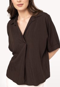 Blusa marrone a maniche corte con colletto, vestibilità rilassata e una sottile texture. Presenta un design drappeggiato con una piega frontale.