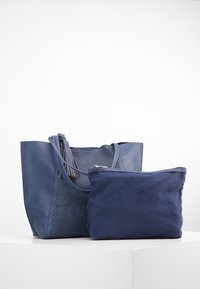 Tmavě modrá kožená taška přes rameno s perforovanou texturou a detaily fringing, doplněná jednobarevnou tmavě modrou zipovou taštičkou.