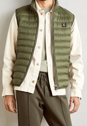 Hombre vistiendo un chaleco acolchado verde oliva sobre una camisa beige con botones y pantalones verdes oliva a juego con cintura ajustable, contra un fondo liso.