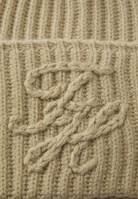 Ulllue i lys beige med ribbestruktur. Har et brodert sammenflettet design i samme farge foran.