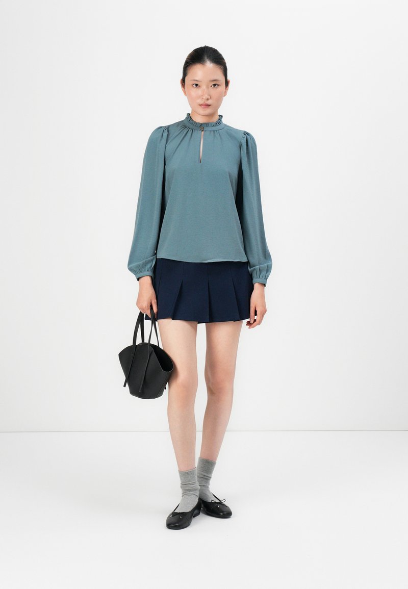 Teal blouse met een gerimpelde halslijn, sleutelgatdetail en lange mouwen. Gecombineerd met een marineblauwe geplooide rok, grijze sokken en zwarte ballerina's.