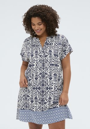 Kobieta z kręconymi włosami, ubrana w wzorzystą sukienkę z krótkim rękawem z granatowymi i białymi kwiatowymi oraz geometrycznymi wzorami, uśmiecha się i patrzy w dół.